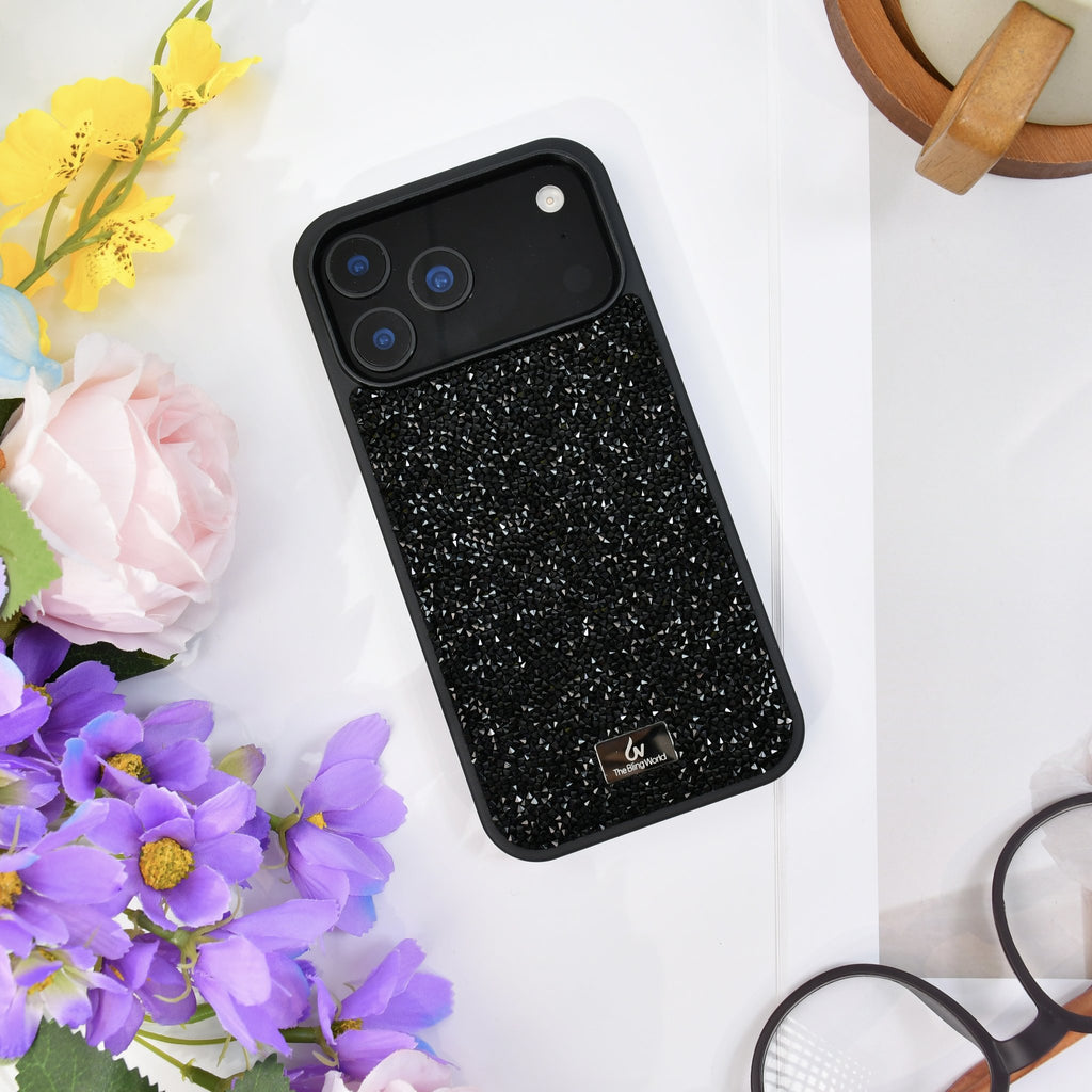 Black rhinestone iPhone case seen from above on white, with yellow and purple flowers and glasses. tbw-rock-black-4