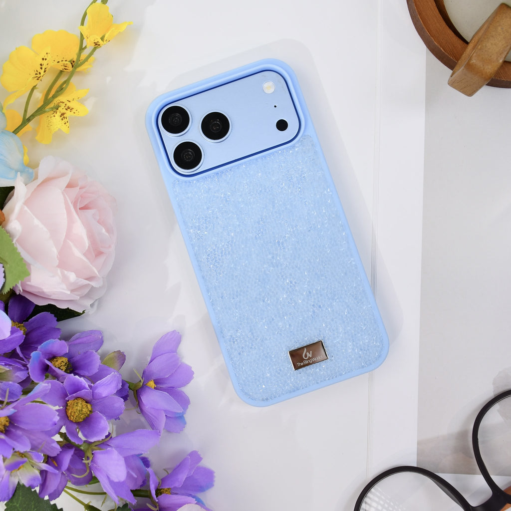 Glacier blue rhinestone iPhone case seen from above on white, with yellow and purple flowers and glasses. tbw-rock-blue-4