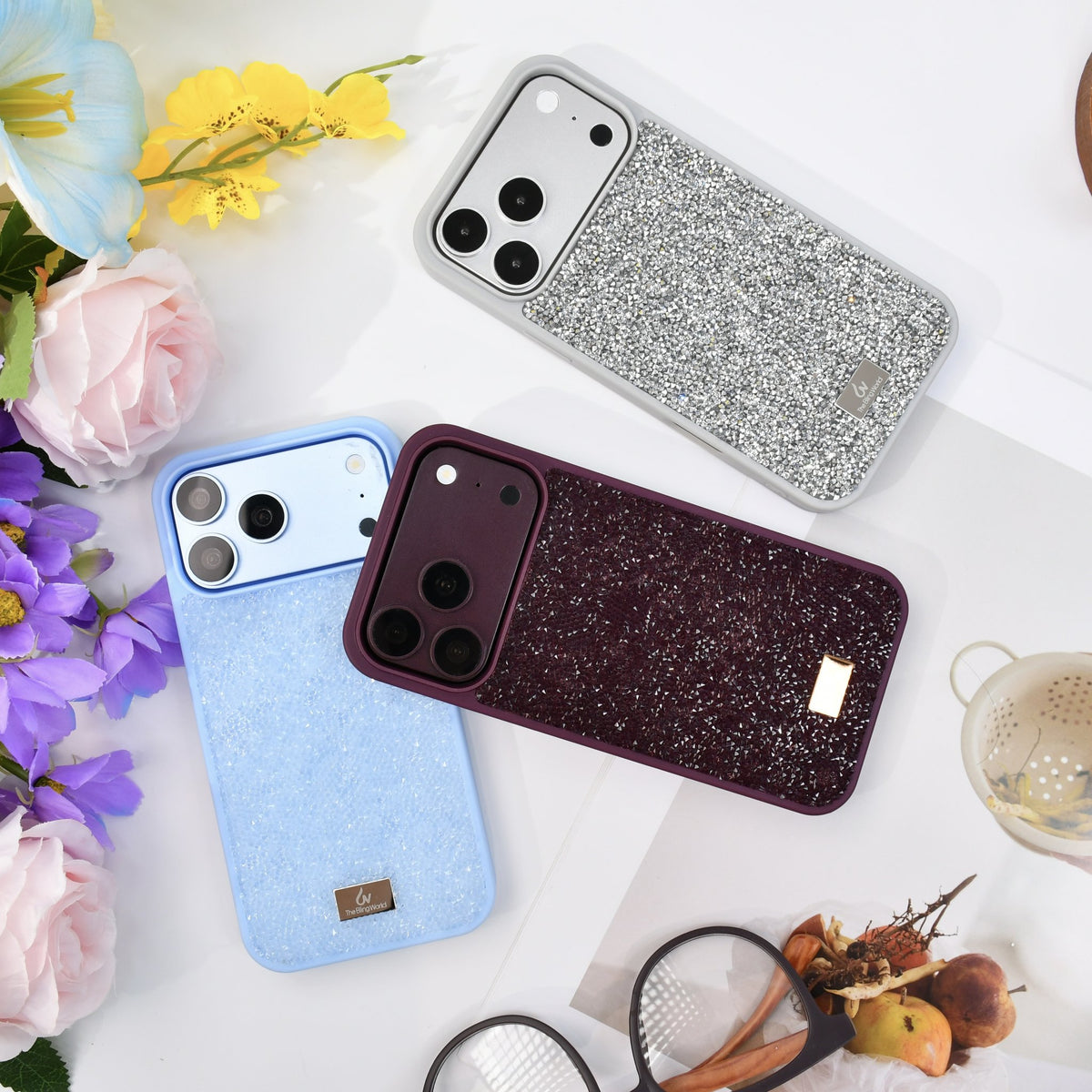 Silver, glacier blue and burgundy rhinestone iPhone cases on white with flowers and a tea strainer. tbw-rock-mix-1